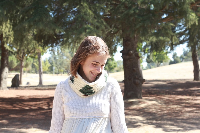 Photo of Claire enjoying the woods and her crocheted Pinewood Tapestry Cowl with a white long-sleeved shirt surrounded by trees while she is looking down